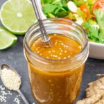 Small mason jar of golden sesame ginger dressing with a bowl of salad and whole ginger on a board.