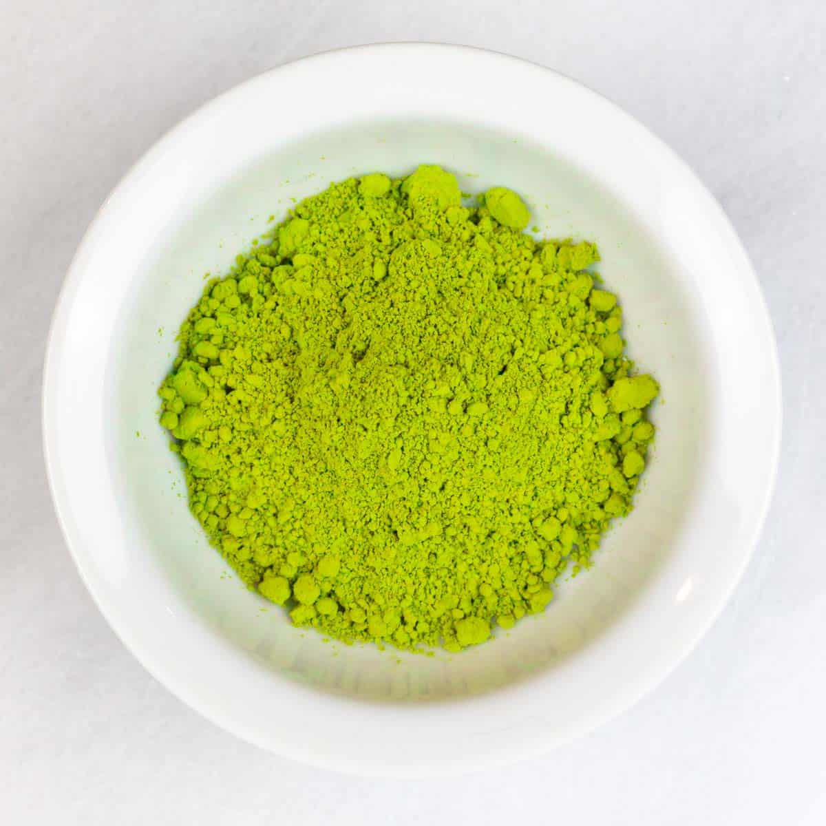 Small white bowl on a white marble board filled with bright green matcha powder.
