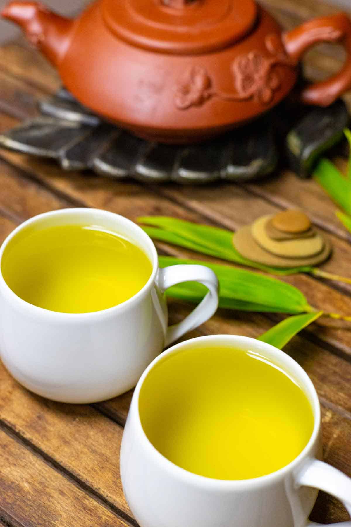 Two small white cups of bamboo tea on a wood slated table with a clay tea pot on a trivet, green leaves and zen rocks.