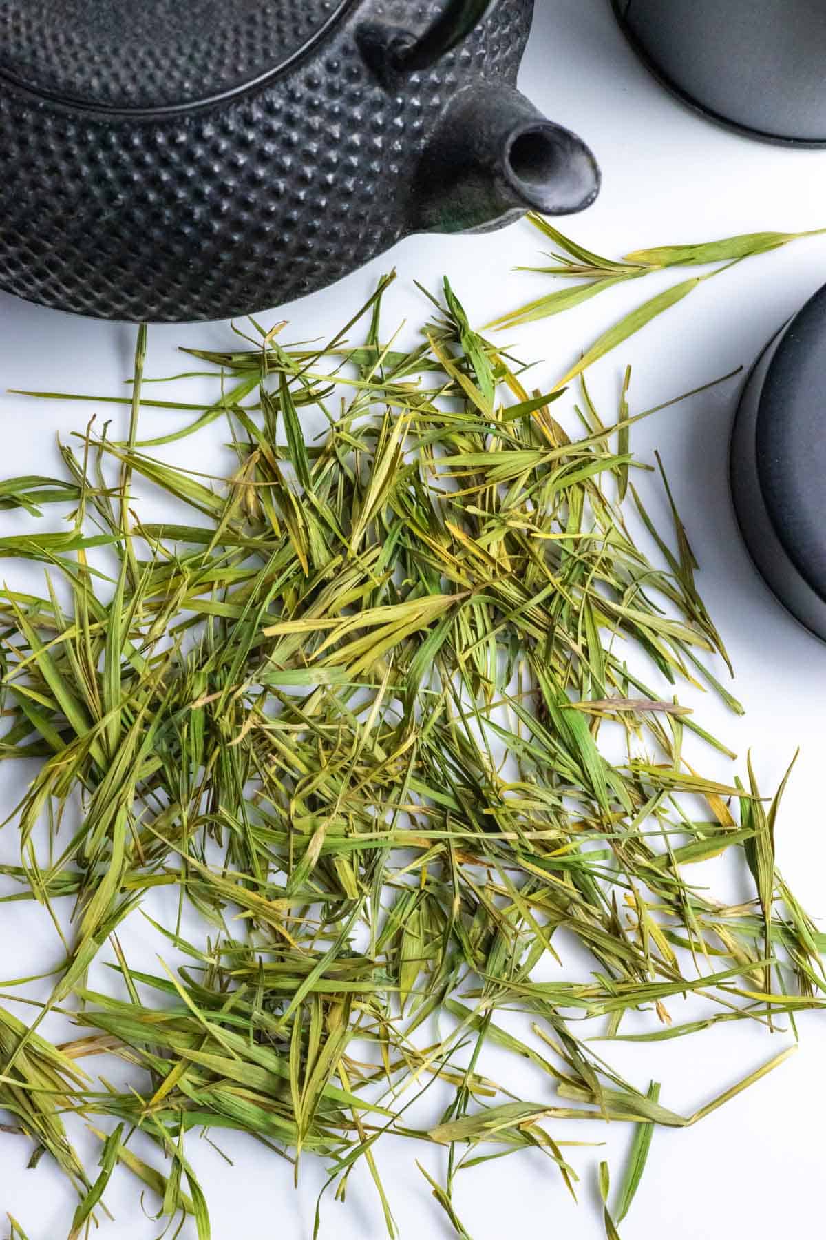 Black iron decorated tea pot on a white board with large pile of green loose leaves of bamboo tea.