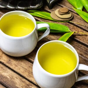 Small white cups of brewed bamboo tea on a wood table with fresh bamboo branch and four small flat stacked rocks in a pyramid.