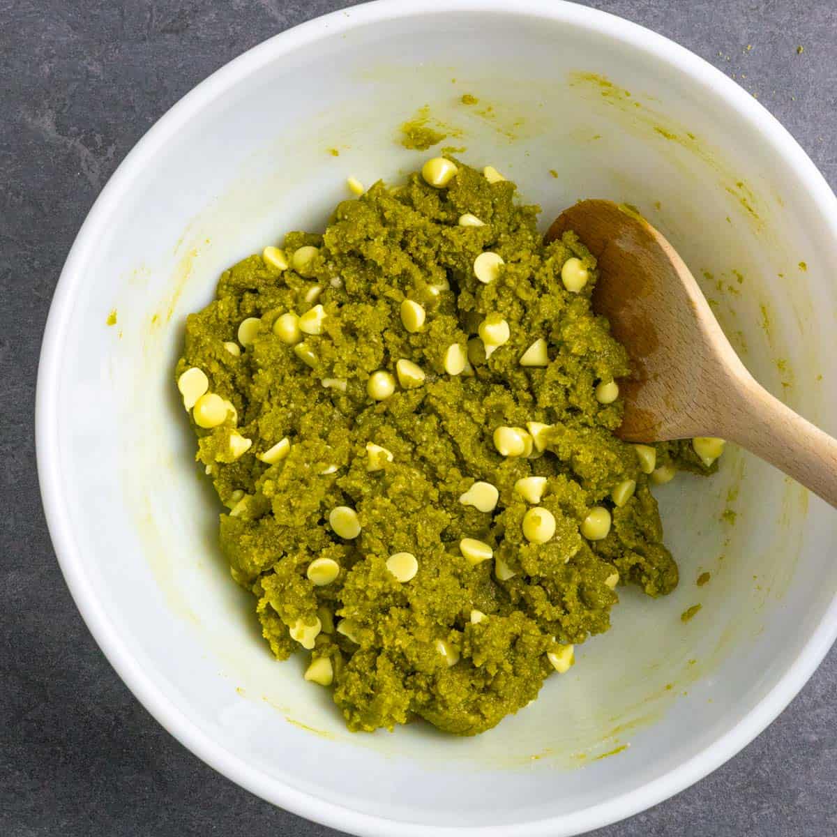 Mixed green cookie dough after adding white chocolate chips and mixing well in a white bowl with a wooden spoon.