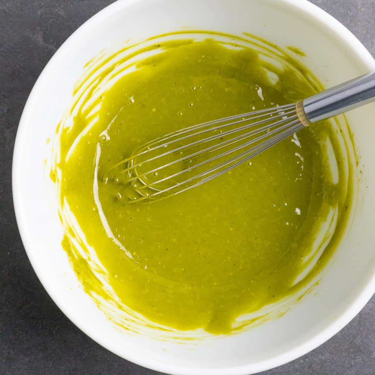 Egg, sweetener, and matcha mixed together in a white mixing bowl with a metal whisk in the bowl.