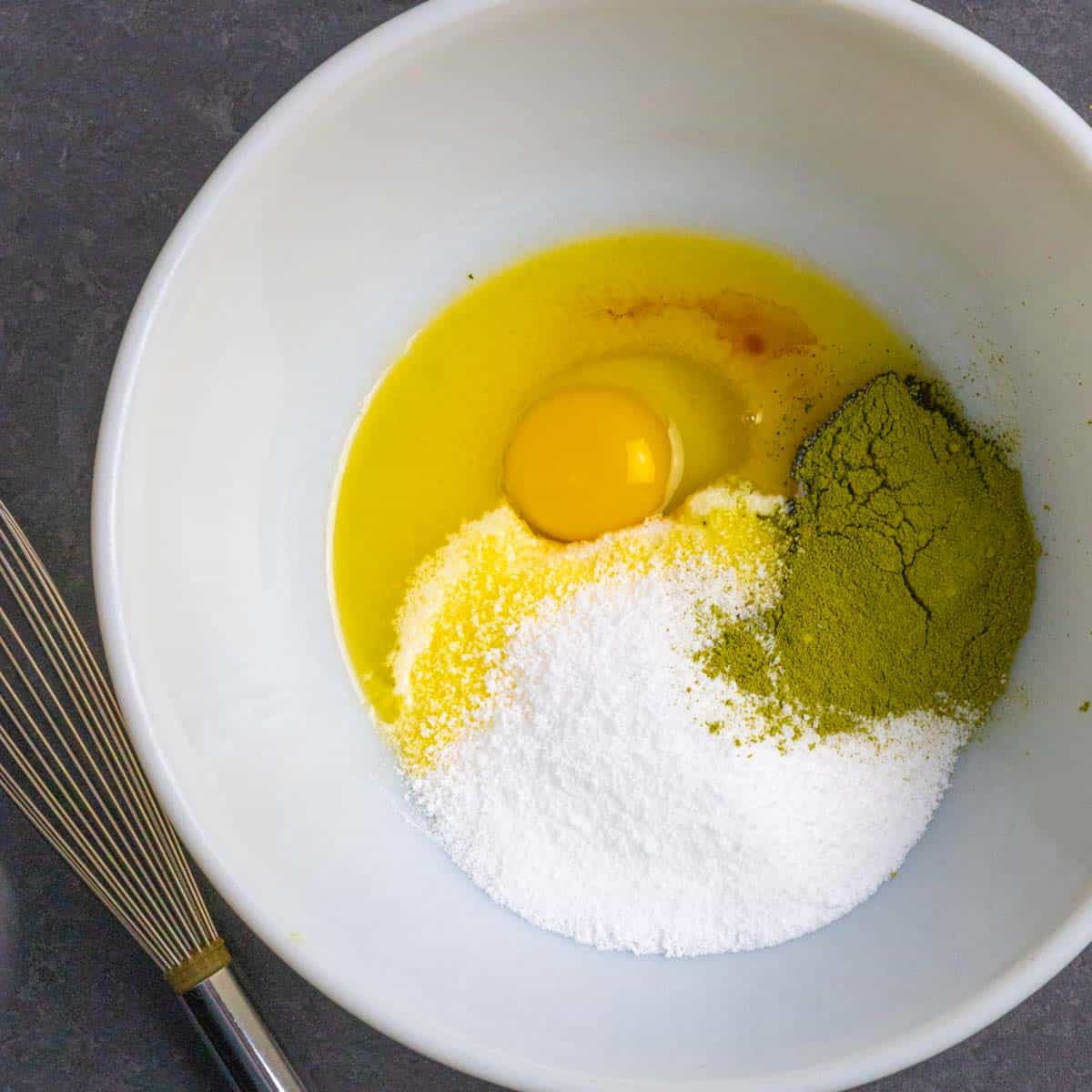 Egg, sweetener, vanilla and matcha added separately to a white mixing bowl with a metal whisk on the side.