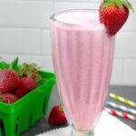 Shake glass of strawberry almond milk smoothie with a berry on the rim and a container of berries next to the glass.