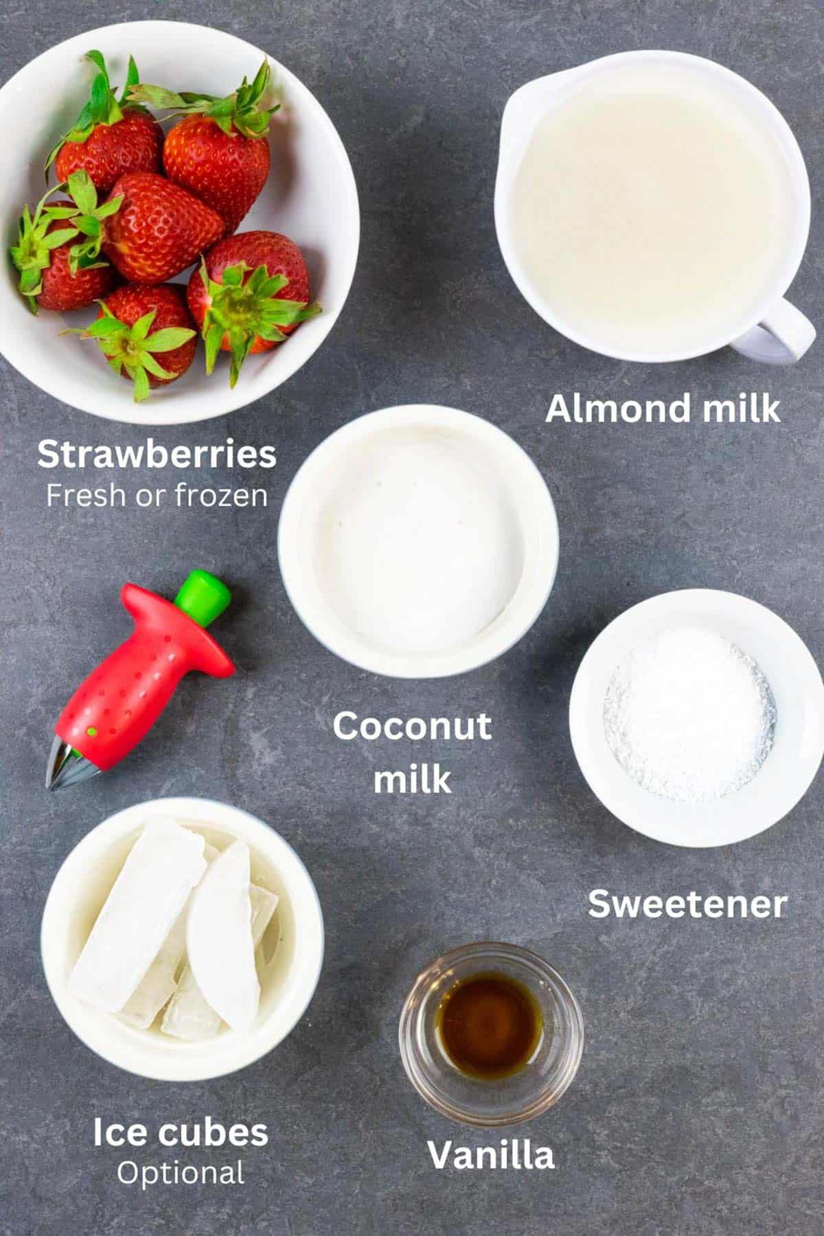 Ingredients for strawberry smoothie in small bowls on a grey board with white text labels.
