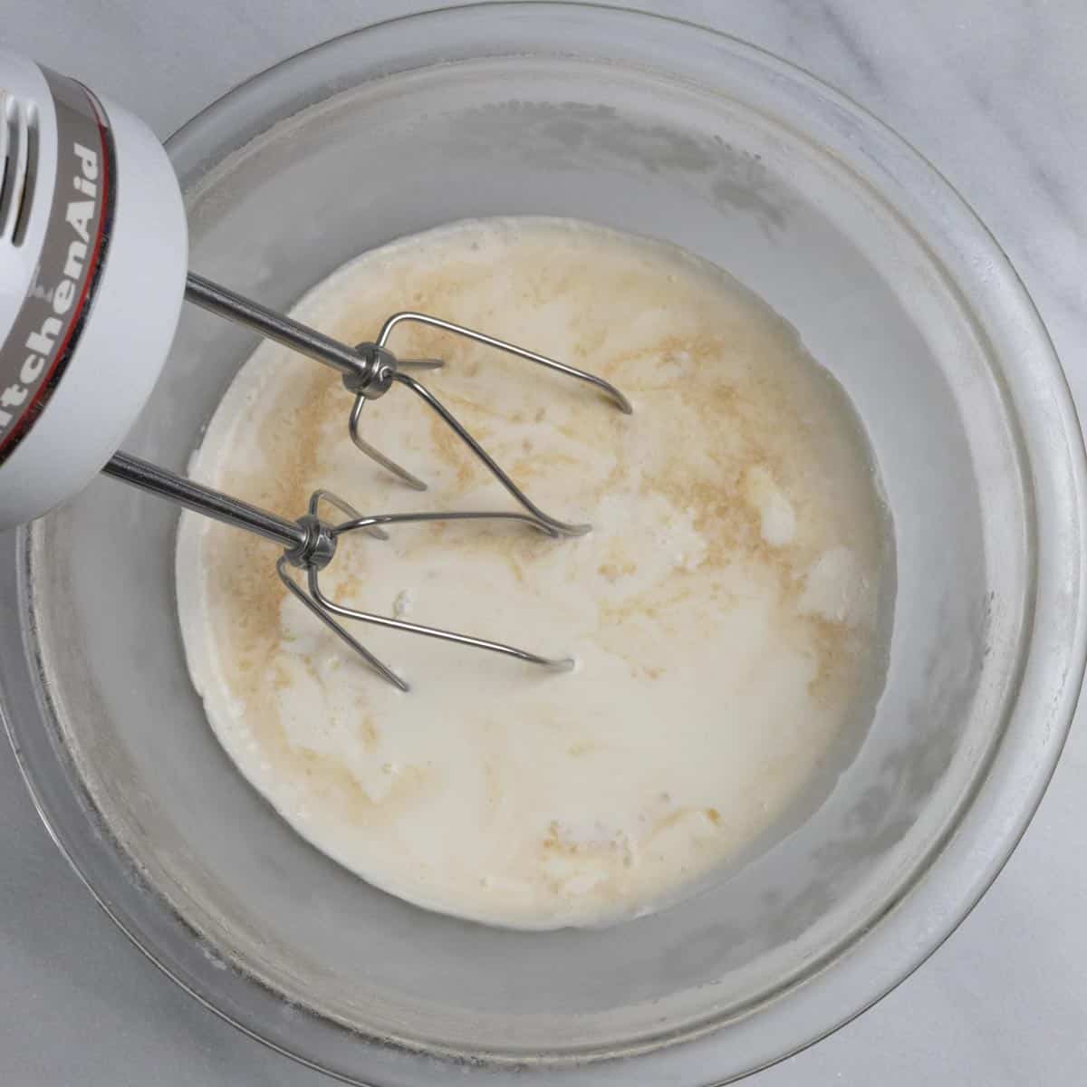 Clear bowl of keto whipped cream ingredients with hand mixer blades in the bowl ready to mix.