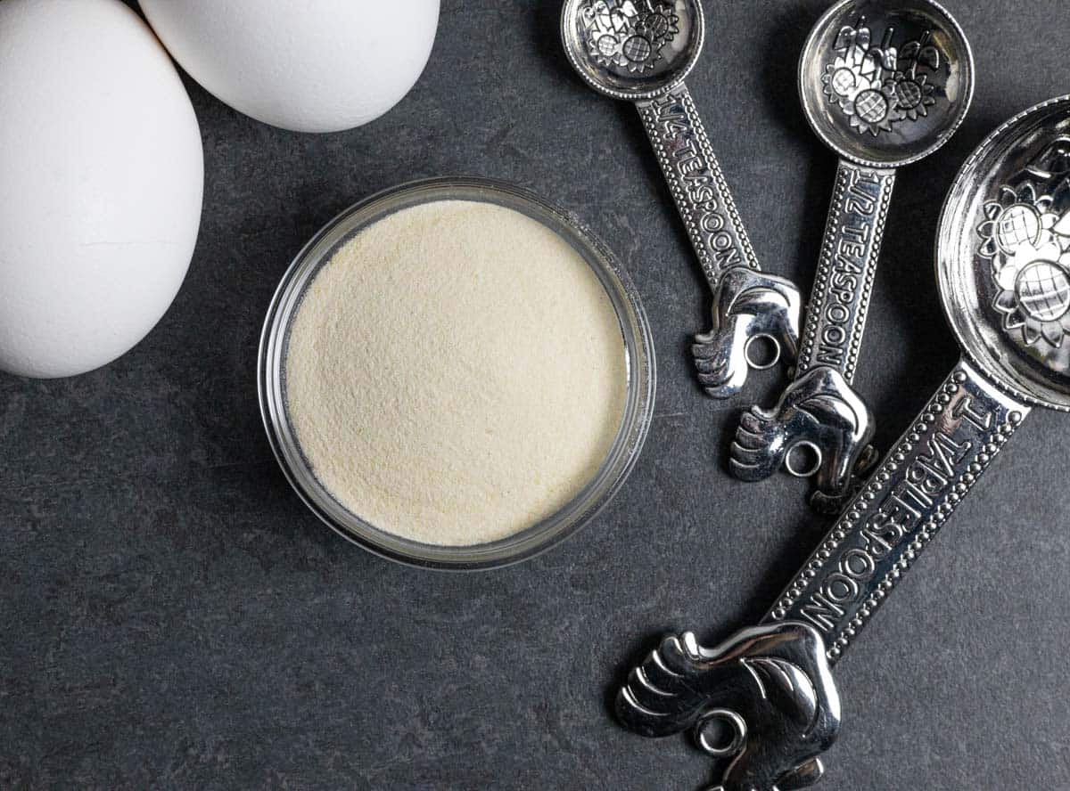 Xanthan gum powder in a small glass bowl with 3 fancy measuring spoons and two whole eggs on the side.