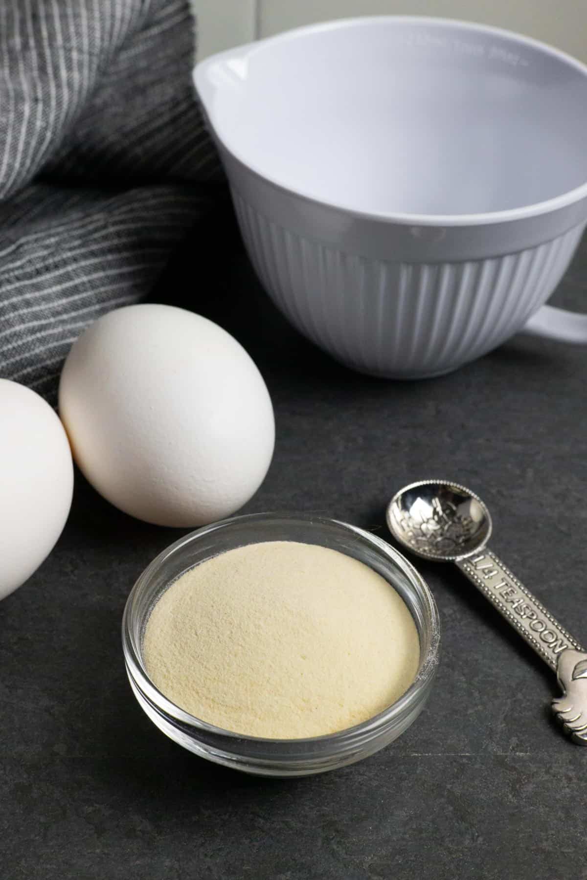 Small bowl of xanthan gum powder with a small white measuring cup, two whole eggs, and a measuring spoon.