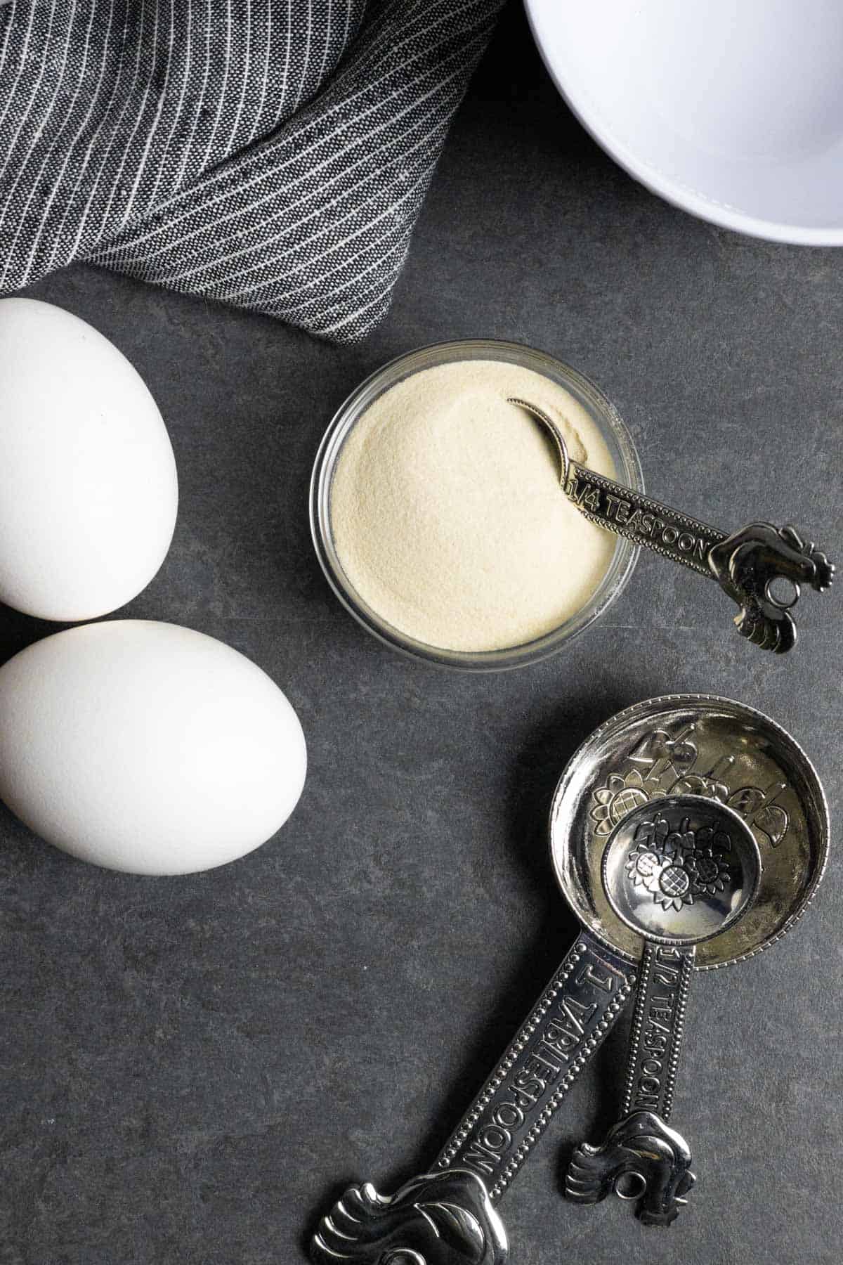 Xanthan gum on a grey board with two whole eggs, three measuring spoons, and a mixing bowl.