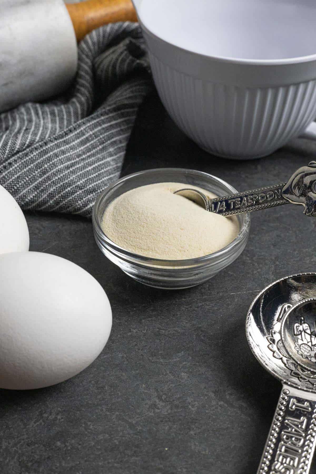 Small glass prep bowl of xanthan gum with measuring spoon on a grey board with 2 whole eggs.