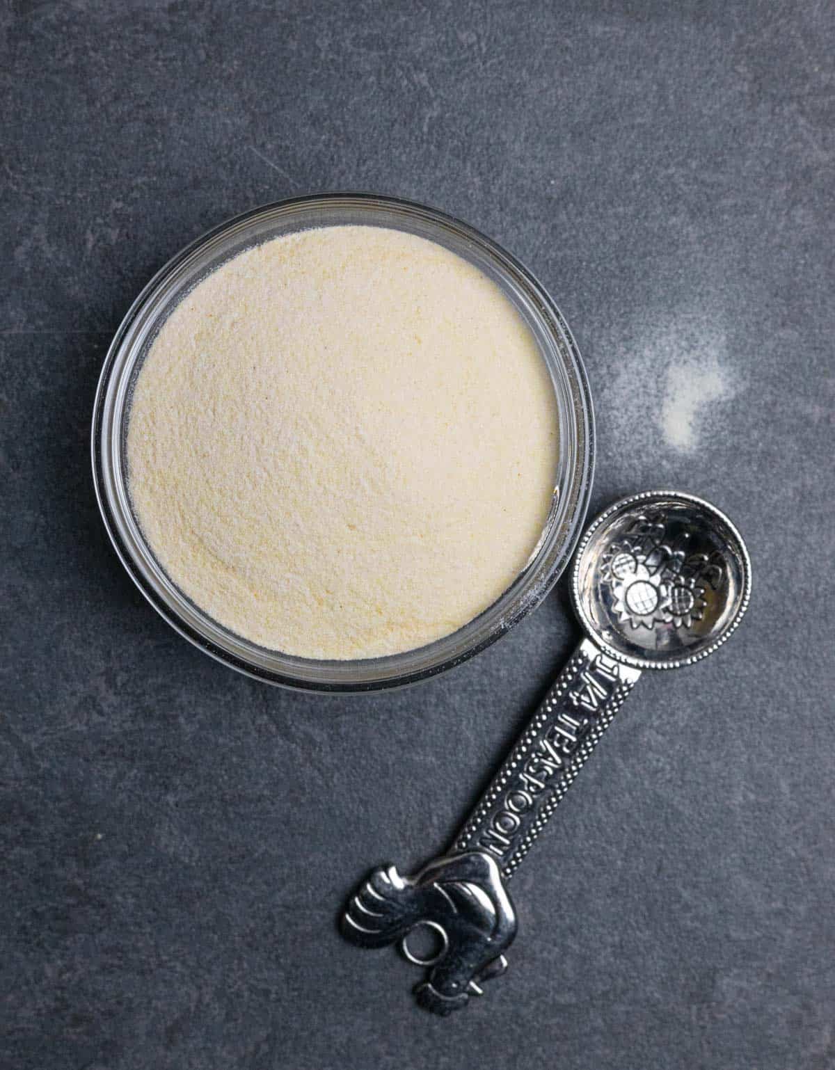 Grey board with a small glass bowl of xanthan gum, a measuring spoon and small sprinkle of powder on the board.