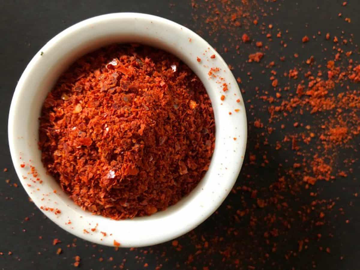 Small white bowl mounded with Aleppo Pepper on a dark board with sprinkles of pepper on the bowl rim and the board.