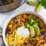 White bowl of chicken taco soup with a spoon, sitting on a marble board next to an instant pot, with lime wedges and cilantro as garnish.