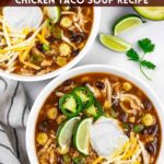 Two white bowls of chicken taco soup topped with shredded cheese, sour cream and lime wedges on a marble board with cilantro, avocado half and lime wedges.