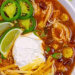 Close look at two thirds of a white bowl of chicken taco soup with toppings of shredded cheese, sliced jalapeno, lime wedges and sour cream.