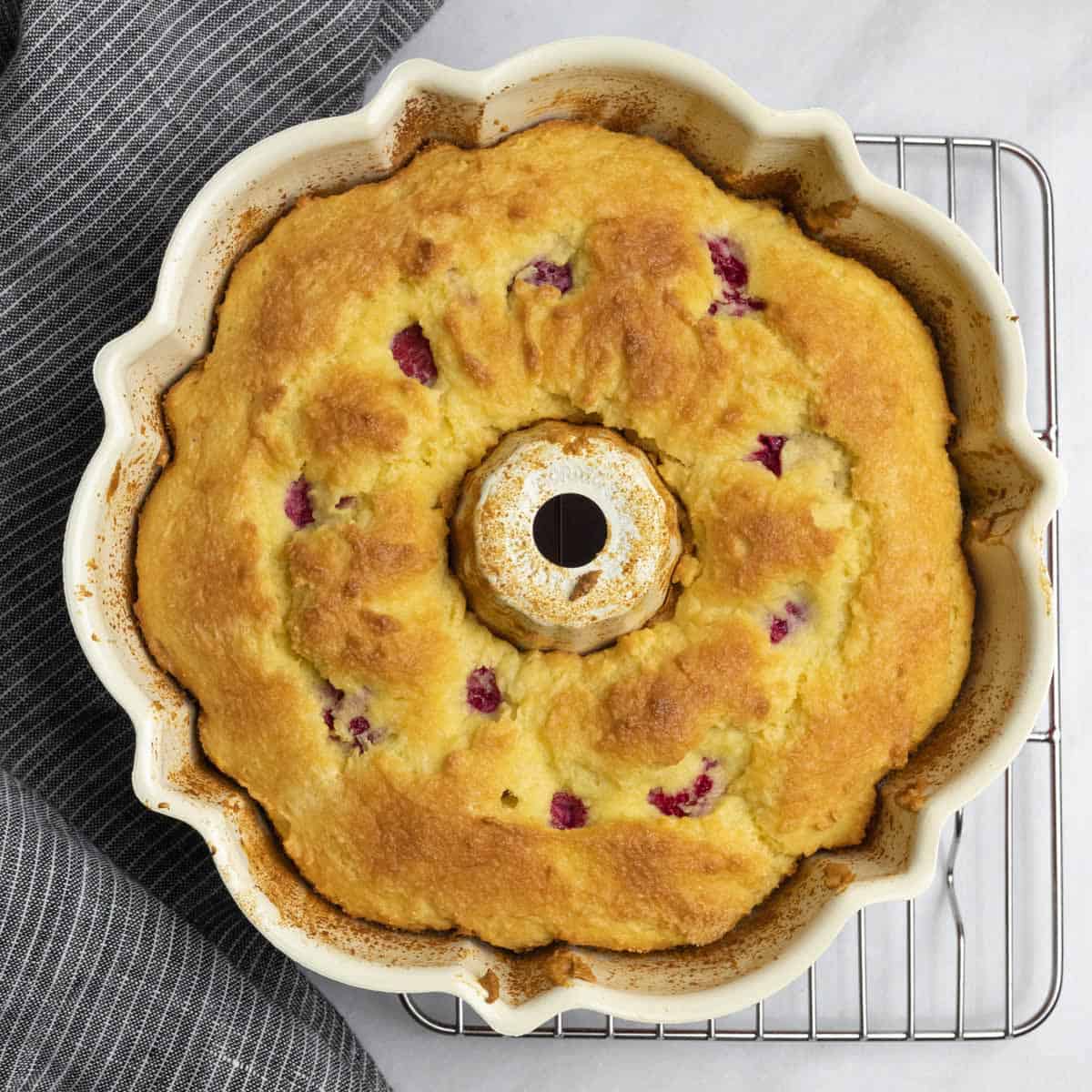 Baked bundt cake in the pan on a cooling rack with golden brown top and raspberries showing through.