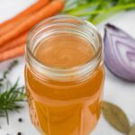 Chicken bone broth in a mason jar with no lid next to a bunch of carrots, a red onion quarter, celery sticks and spices.