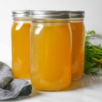 Three quart jars of chicken bone broth, two behind and one in front with a grey napkin in front and a carrot bunch behind.