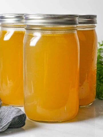 Three quart jars of chicken bone broth, two behind and one in front with a grey napkin in front and a carrot bunch behind.