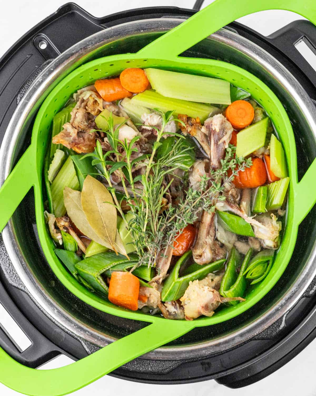 Looking into Instant Pot with a lime green silicone strainer inside, full of raw vegetables, herbs and chicken bones for broth.