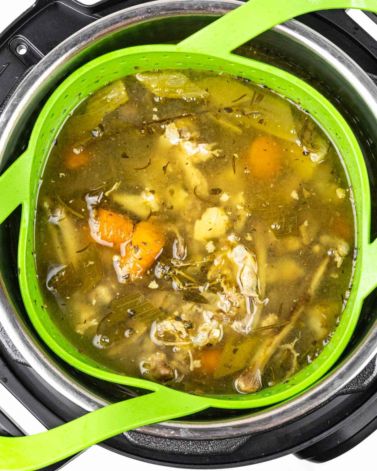 Looking into a full Instant Pot of cooked chicken bone broth showing vegetables, herbs and golden broth.