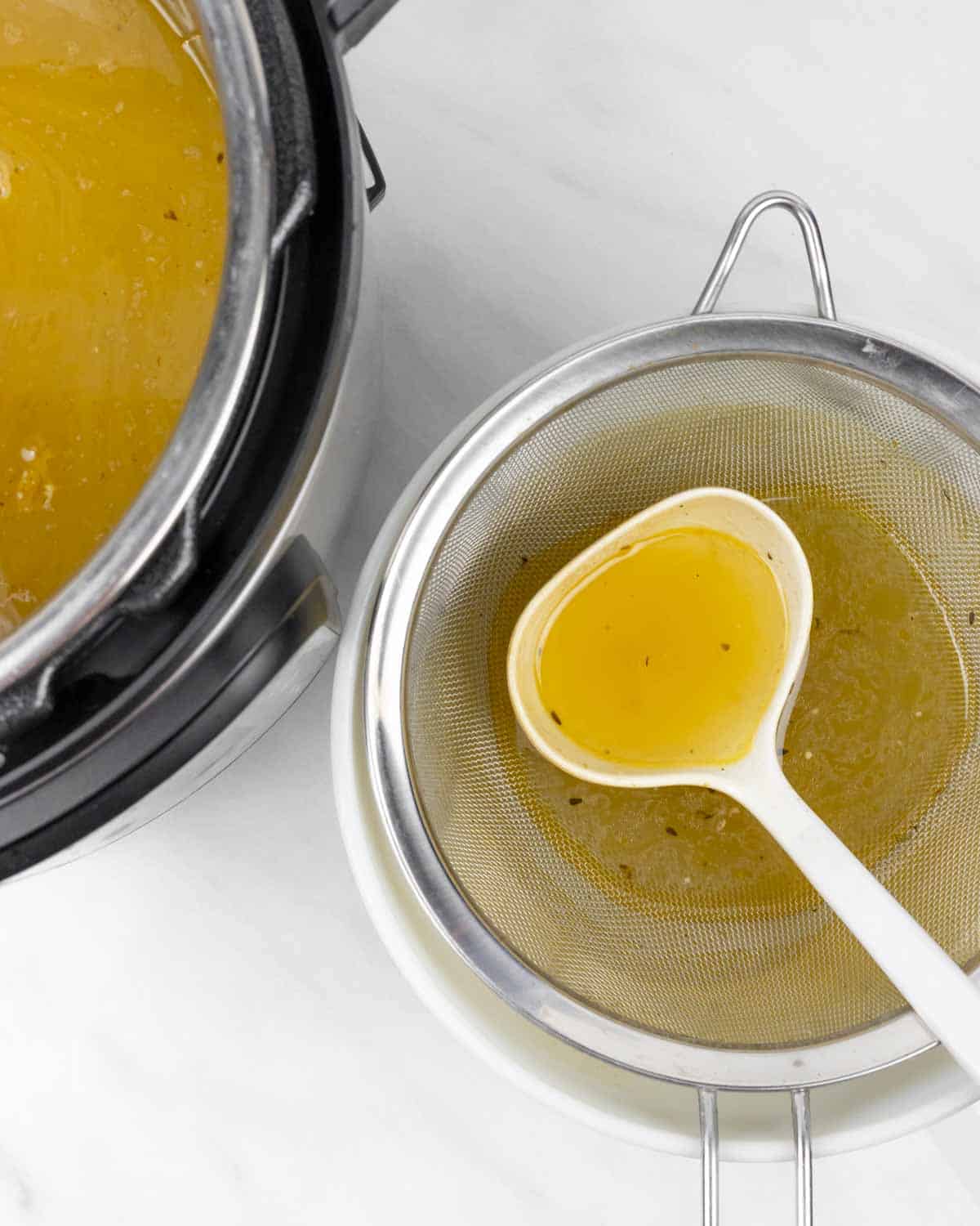 Ladel and strainer in a large bowl of broth sitting next to an Instant Pot with the rest of the broth.