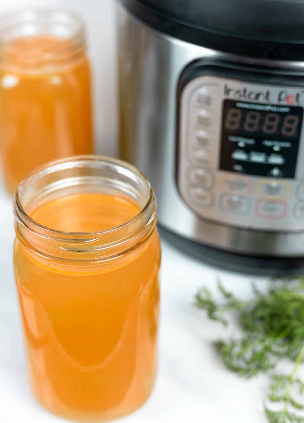 Open quart jars of chicken broth in front of an Instant Pot with herbs sprinkled in front.