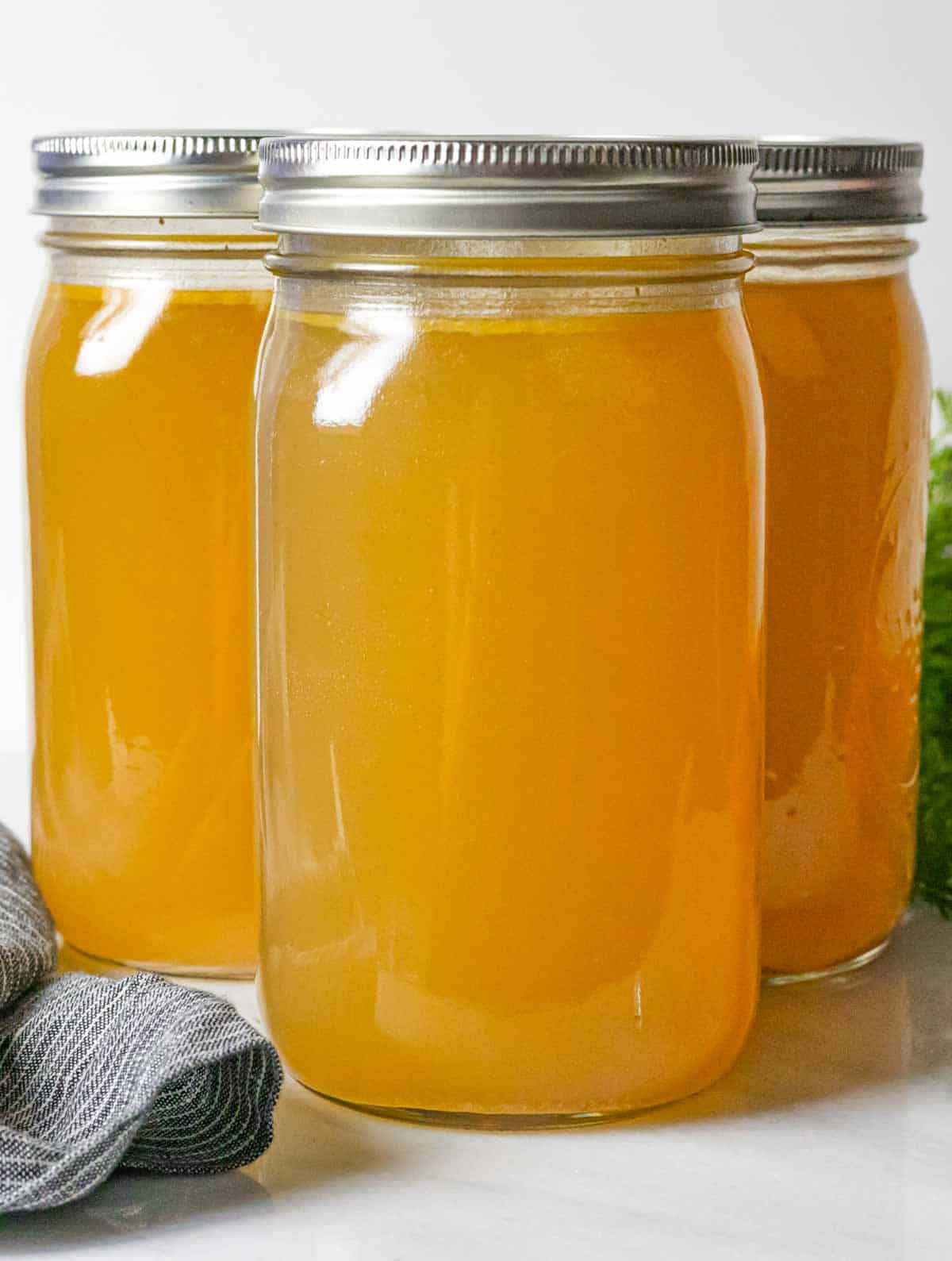 Three quart jars of finished chicken bone broth, two behind and one in front with a grey napkin in front and a carrot bunch behind.