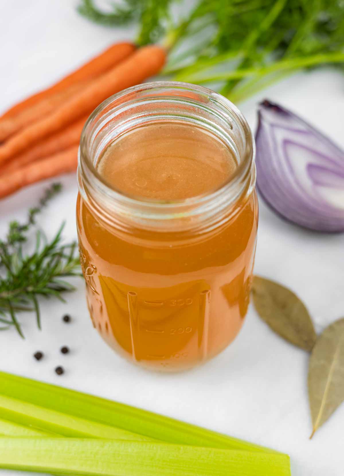 Chicken bone broth in a mason jar with no lid next to a bunch of carrots, a red onion quarter, celery sticks and spices.