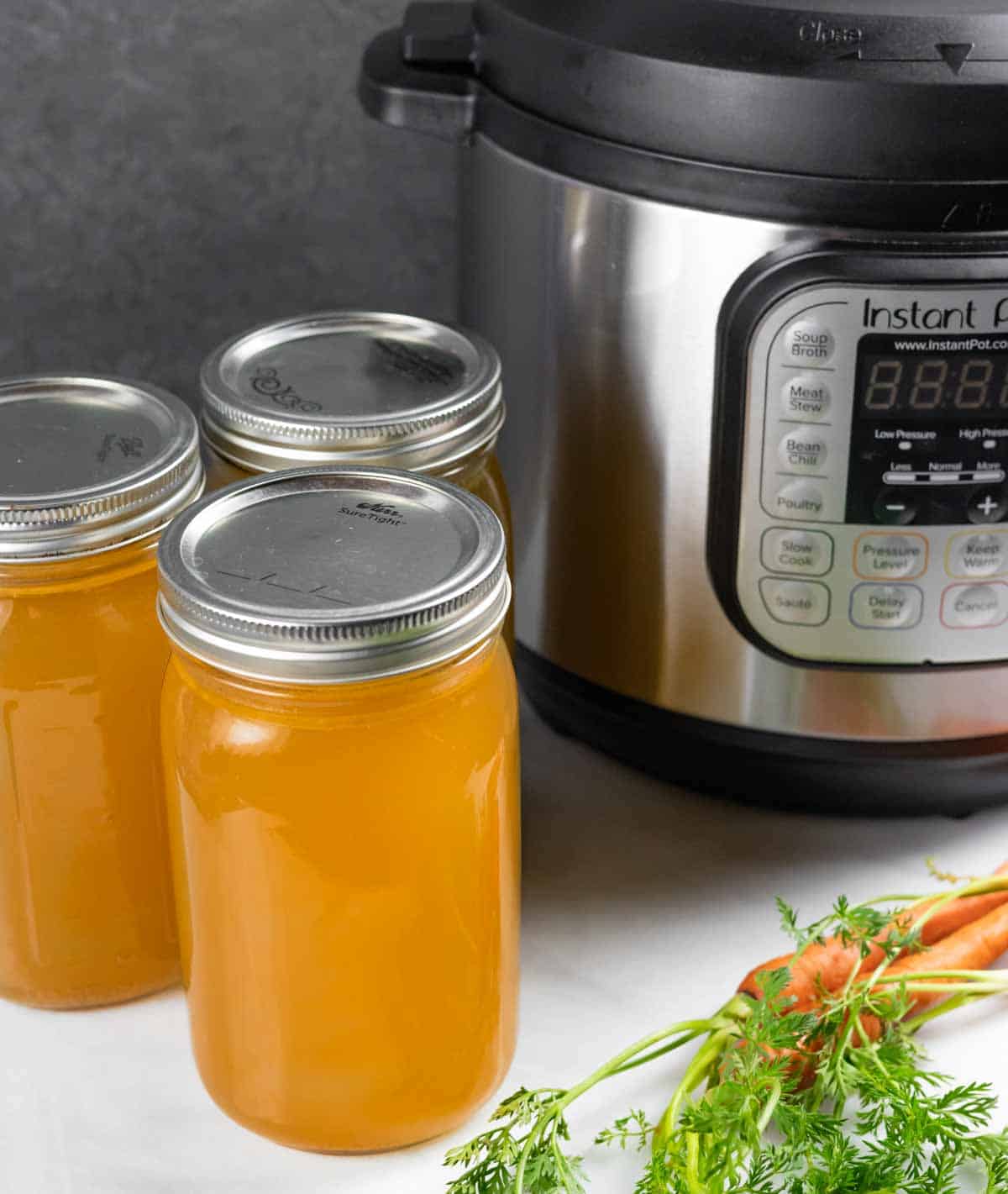 Three lidded quart jars of chicken bone broth next to an Instant pot with a small bunch of orange carrots with tops in front.