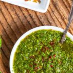 White bowl of Mexican chimichurri sauce sprinkled with Aleppo pepper with a spoon in the bowl on a textured clay surface.