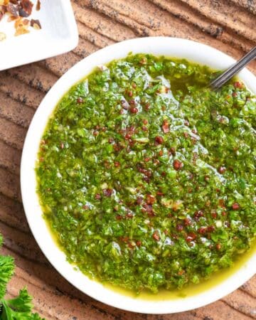 White bowl of chimichurri sauce with a spoon in it on a clay textured plate with pepper flakes and parsley on the side.