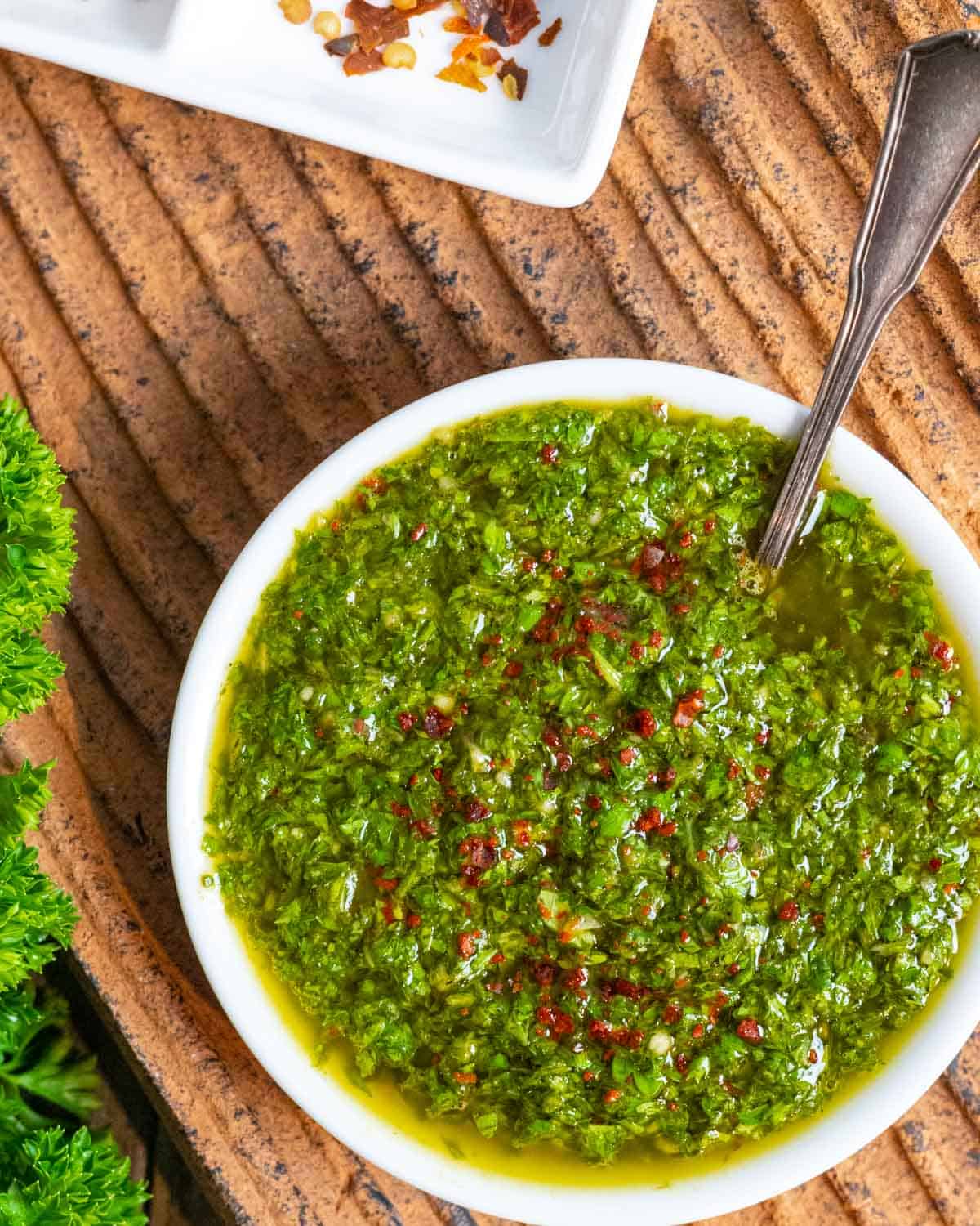 White bowl of Mexican chimichurri sauce sprinkled with Aleppo pepper with a spoon in the bowl on a textured clay surface.