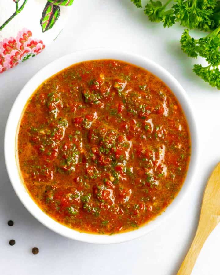 Bowl of red chimichurri sauce on a white board with a small wood spoon next to it, a decorative towel, and parsley.