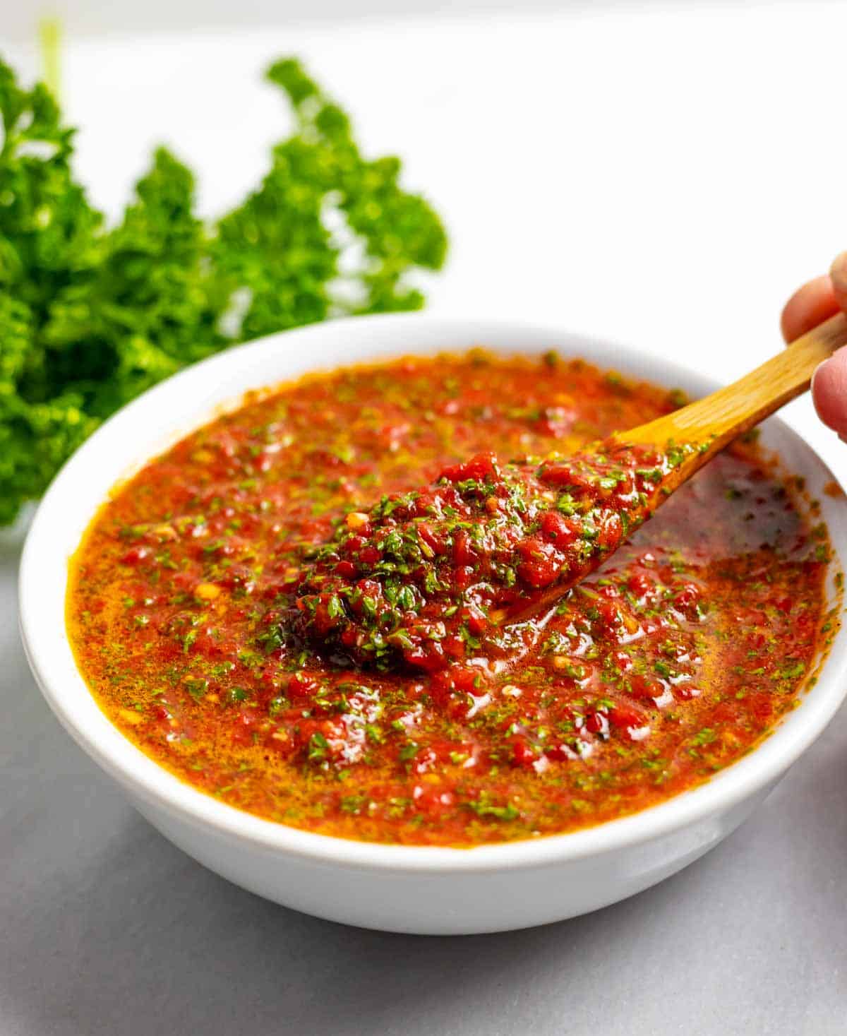 White bowl of chimichurri rojo with a hand lifting a small wood spoon full of delicious red and green sauce.
