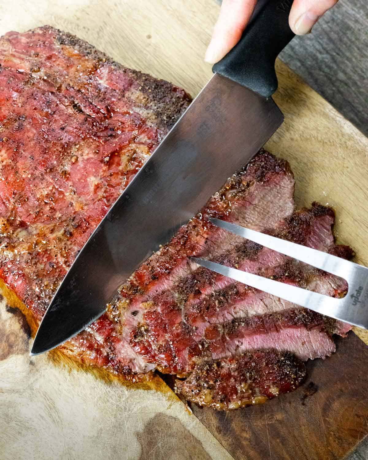 Partially sliced smoked flank on a board with carving fork and knife slicing a new thin slice.