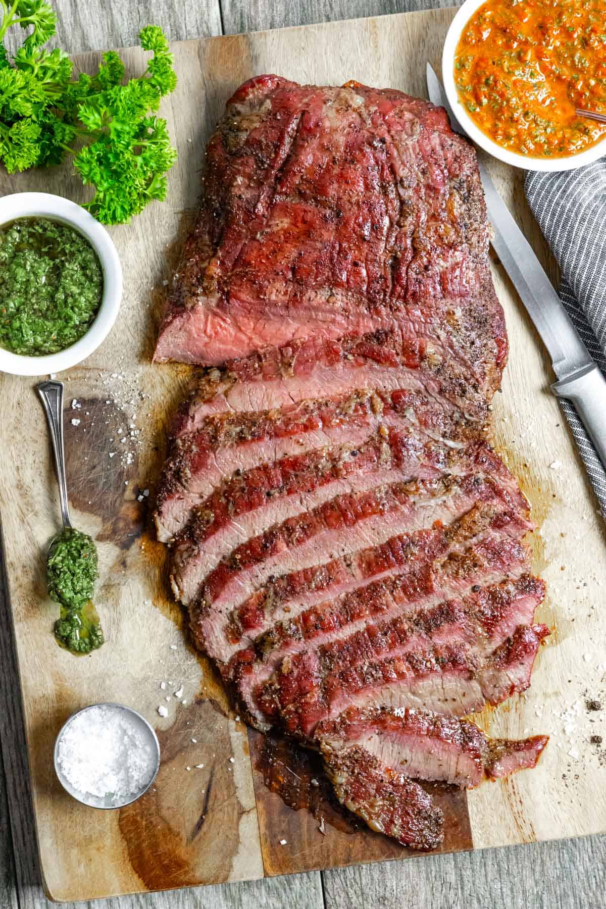 Smoked flank steak with half sliced on a board with small white bowls of green and red chimichurri sauce.