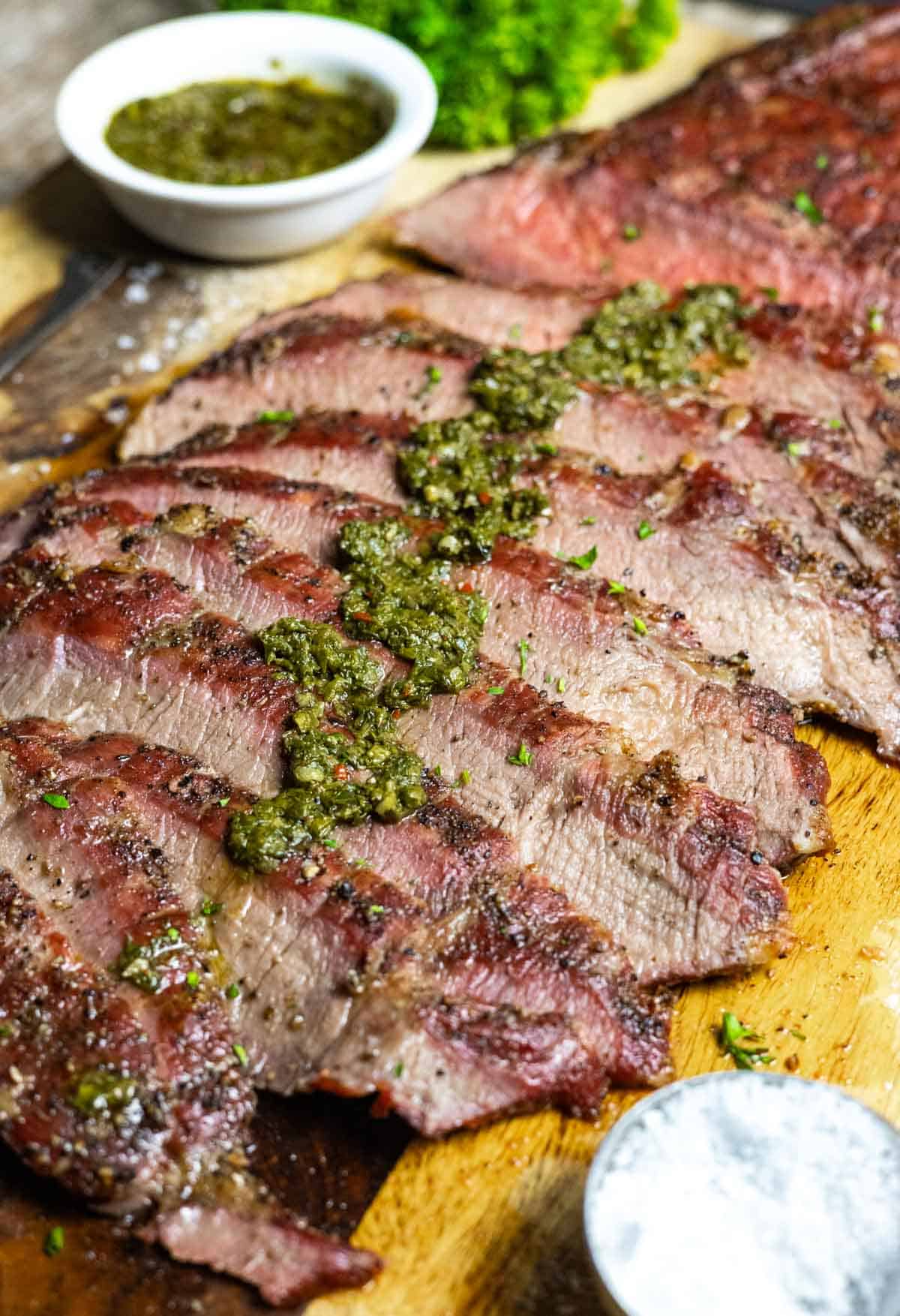 Layered slices of steak on a board with chimichurri verde down the center of slices with a small bowl of sauce and salt.