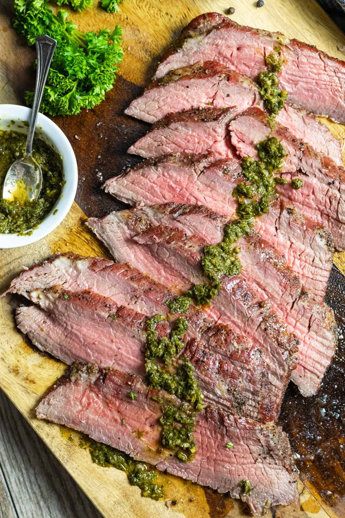 Nine thin slices of smoked flank steak layered over each other on a board with green chimichurri sauce spread down the center of each slice.