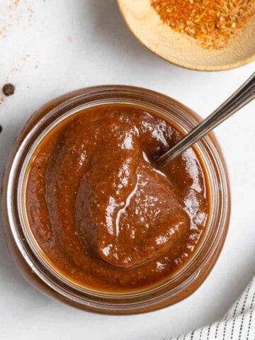 Top down look into a jar of low-FODMAP barbeque sauce with a spoon in the jar and spices surrounding.