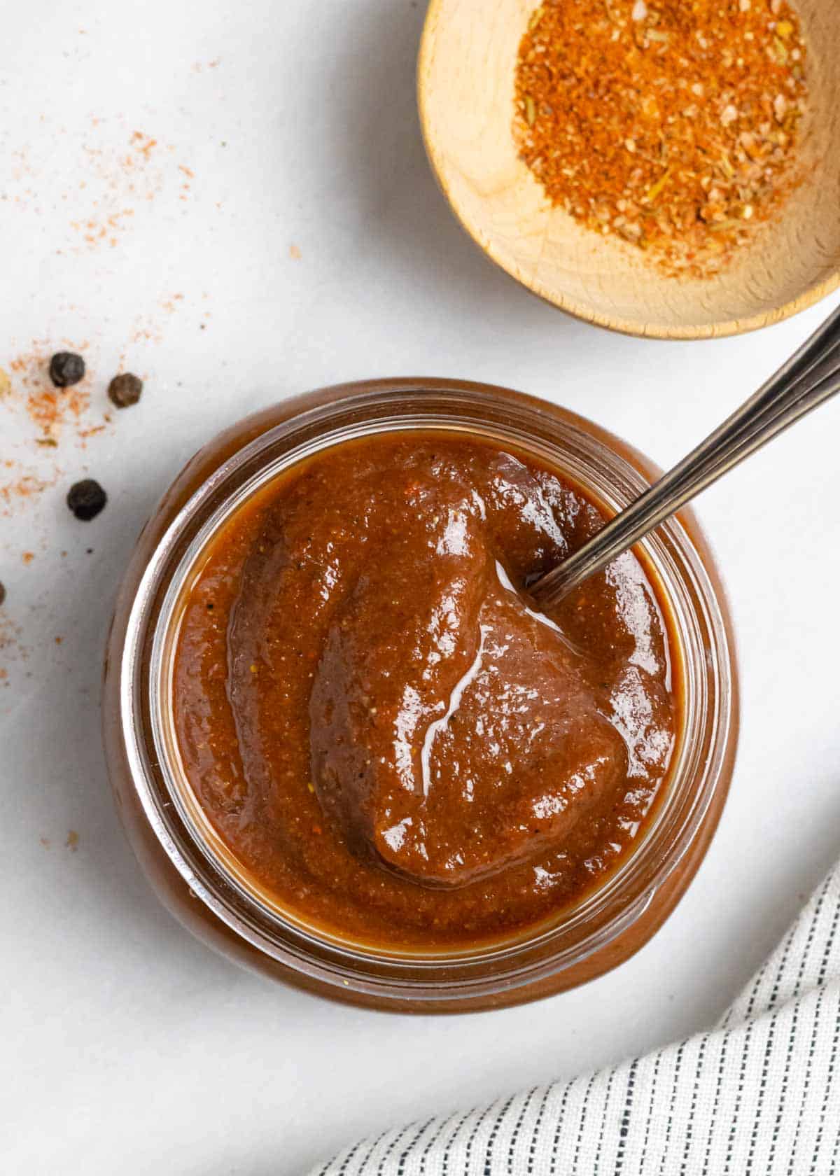 Looking into top of a glass jar of rich, red, low fodmap barbeque sauce next to a small wood bowl of spices.