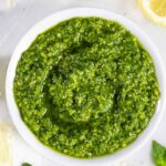 Pesto without pine nuts in a ramekin on a white board with lemon slices, basil leaves and peppercorns.