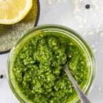 Looking down into jar of pesto with small silver spoon in it with lemon halves on the side.