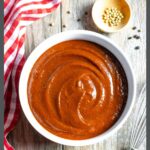 White bowl of no cook BBQ sauce on a board with a red checkered towel and small bowl of peppercorns.