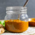 Tikka masala paste in a small glass mason jar with a spoon in the jar and a bowl of spice and a ginger root next to it.