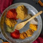 Small piles of tikka spices in a circle on a pewter plate with a small wooden spoon in the center.