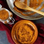 Tikka spice in a small wood bowl on a red scarf with a small spice jar of spice next to it.