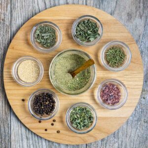 Jar of all purpose seasoning blend on a round board surrounded by small bowls of whole herbs.