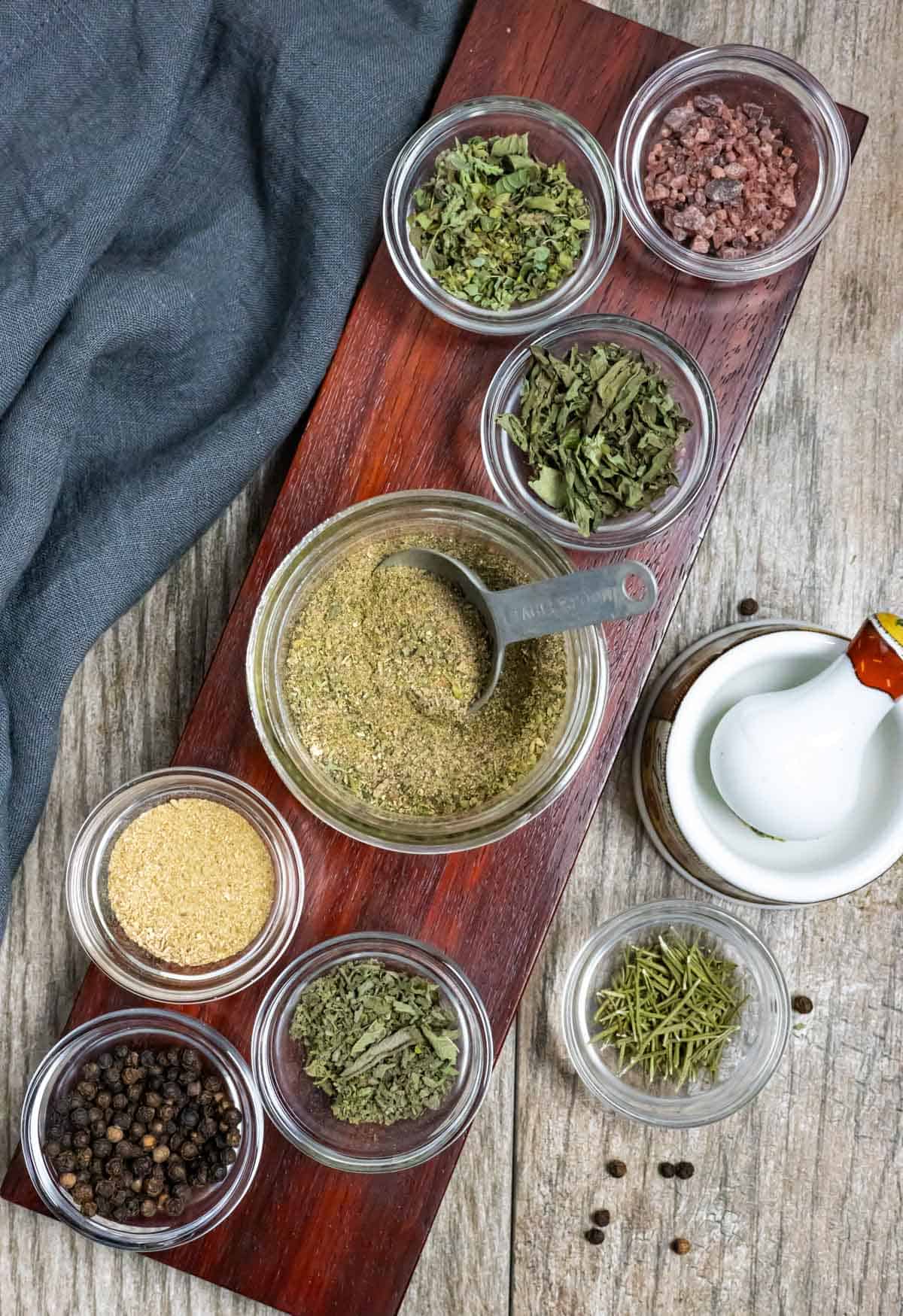 Angled dark narrow red-brown board with small glass bowls of whole spices and glass jar of ground spice blend in the center.